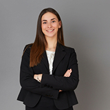 A woman in a black blazer and white blouse stands confidently with folded arms against a gray background, conveying professionalism and assurance.