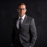 A man in a dark suit and tie stands confidently against a black background. His hands are in his pockets as he gazes at the camera, exuding professionalism.
