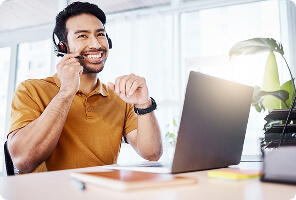 Happy male customer service representative with headset speaking while using a laptop at desk