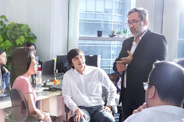 A man in a suit speaks to a group in an office, conveying a focused and collaborative atmosphere. Colleagues listen attentively, surrounded by desks and computers.