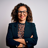 A woman with curly hair and glasses smiles confidently, wearing a black blazer over a leopard print top. Her arms are crossed, exuding a professional vibe.