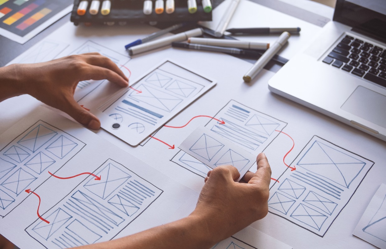 Designer hands arranging paper wireframes and website UI sketches on a desk next to a laptop and markers.