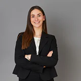 A woman in a black blazer and white blouse stands confidently with folded arms against a gray background, conveying professionalism and assurance.