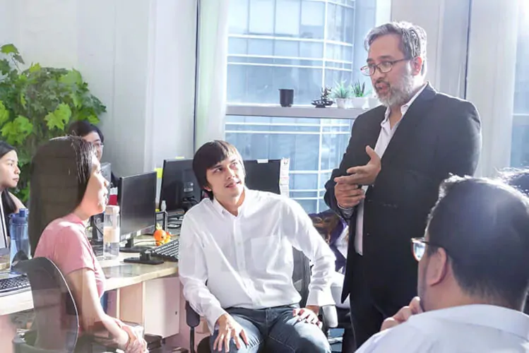 A man in a suit speaks to a group in an office, conveying a focused and collaborative atmosphere. Colleagues listen attentively, surrounded by desks and computers.