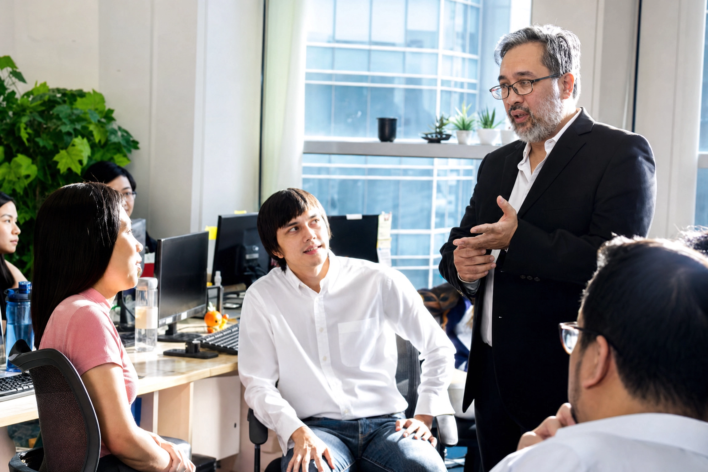 A man in a suit speaks to a group in an office, conveying a focused and collaborative atmosphere. Colleagues listen attentively, surrounded by desks and computers.